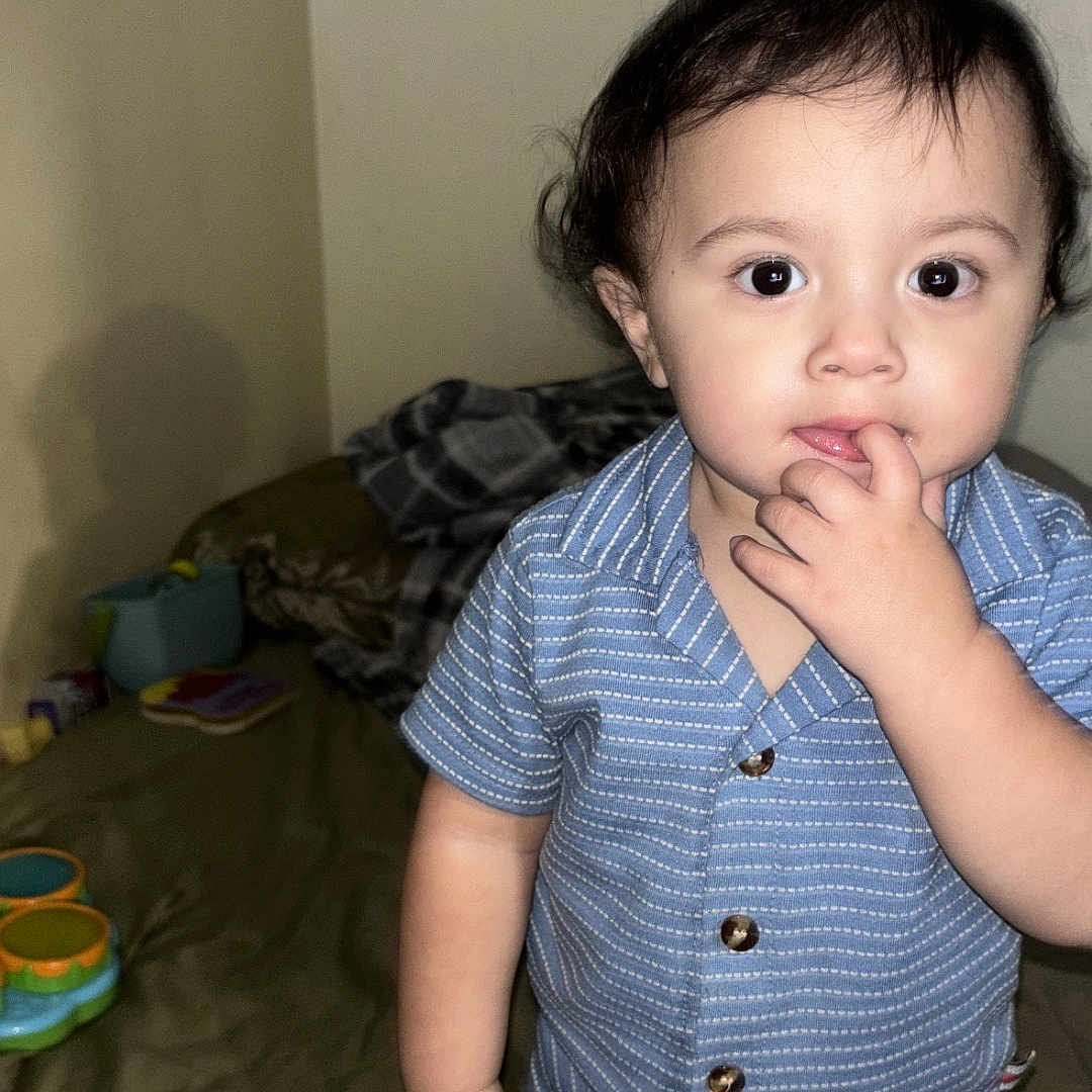 Jayceon is registered to the contest to win money with this photo: bed, bedroom, blanket, blue_shirt, child, clothing, curious, expression, face, fingers, hand, home, indoor, person, portrait, shorts, standing, toddler, toys, young_child