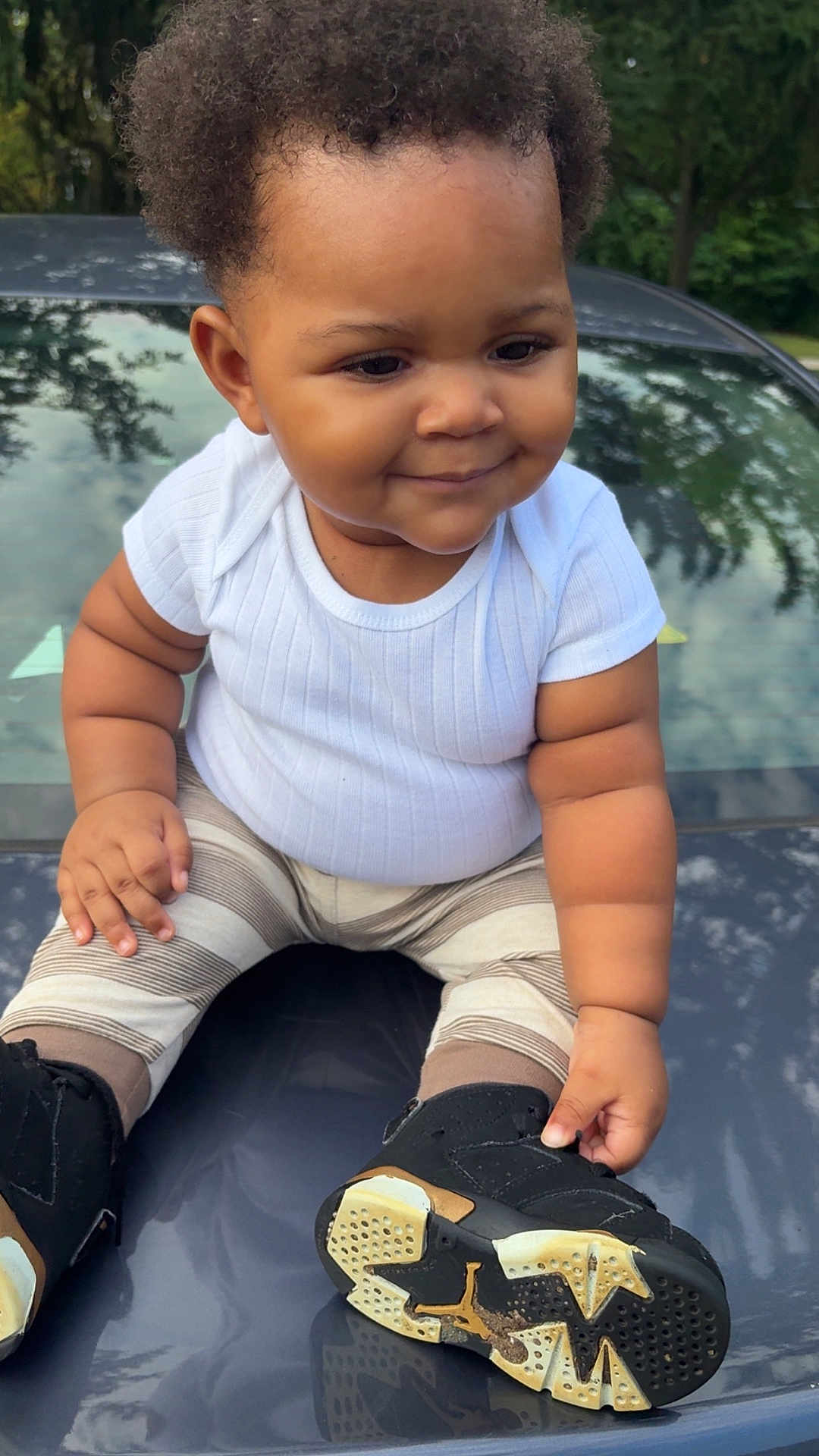 Desmoni joined the competition — help win amazing prizes! toddler, child, baby, white_shirt, striped_pants, black_shoes, car_hood, outdoor, trees, reflection, smiling, cute, sitting, hand, footwear, curly_hair, chubby_cheeks, daylight, casual_clothing, playful