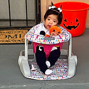Maggie is registered to the contest to win money with this photo: baby, infant, baby_seat, plush_toy, pumpkin, halloween, jack_o_lantern, door_mat, porch, pigtails, white_bows, black_pants, white_socks, pacifier, front_door, concrete, colorful_pattern, cute, festive, outdoor