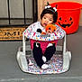 baby, infant, baby_seat, plush_toy, pumpkin, halloween, jack_o_lantern, door_mat, porch, pigtails, white_bows, black_pants, white_socks, pacifier, front_door, concrete, colorful_pattern, cute, festive, outdoor