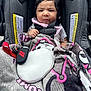 baby, child, car_seat, blanket, pink_bows, clothing, face, person, seatbelt, indoor, cute, infant, expression, hand, keychain, black_hair, fabric, sitting, portrait, warning_label