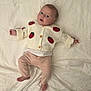 Amelia is registered to the contest to win money with this photo: baby, infant, child, person, face, eyes, sweater, strawberry_pattern, clothing, pants, bed, bedding, white_sheet, lying_down, cute, expression, skin, foot, hand, indoor