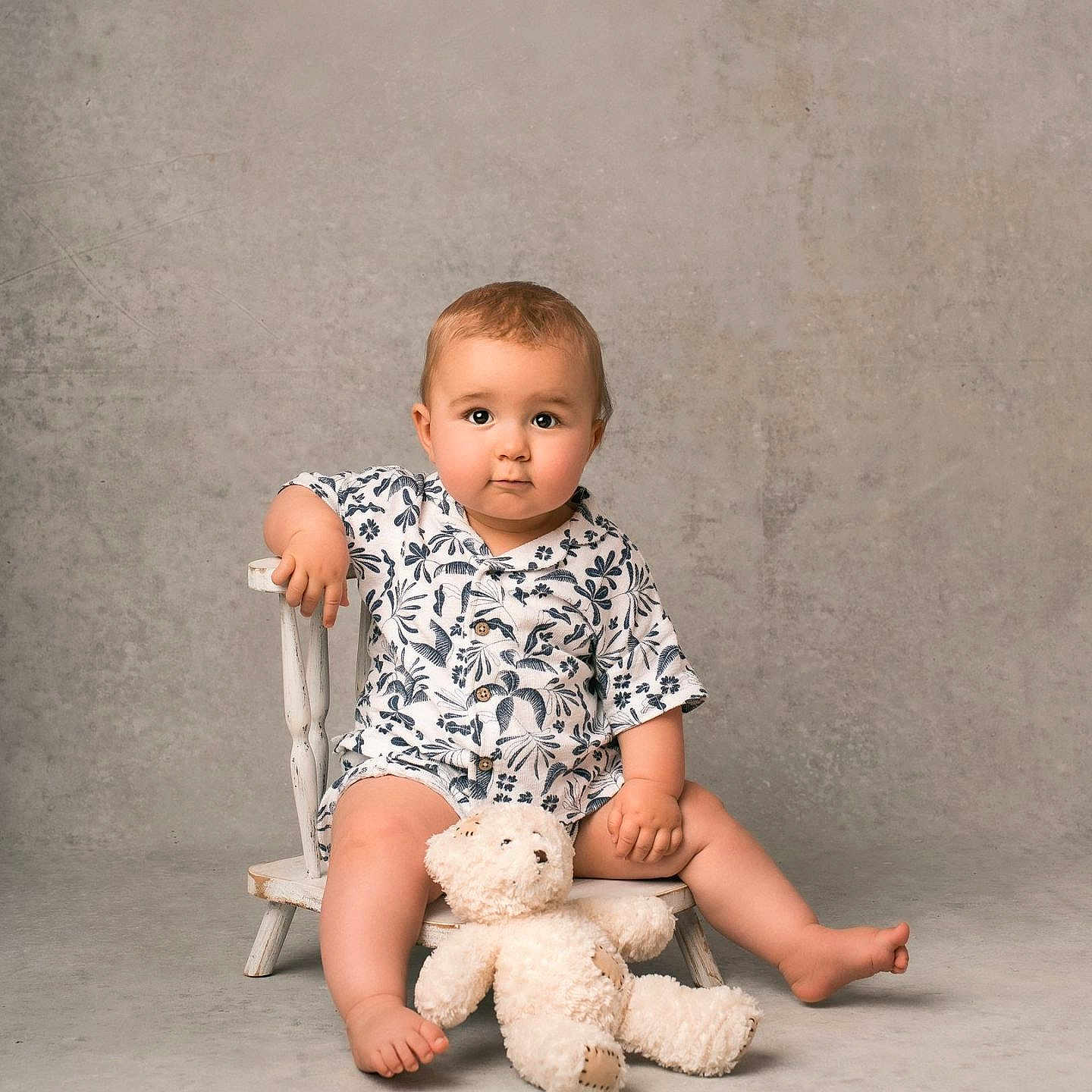 Emery a rejoint le concours — aidez-le/la à gagner de superbes lots ! toddler, child, baby, sitting, chair, teddy_bear, plush_toy, romper, floral_pattern, barefoot, indoor, portrait, cute, calm, curious, studio, neutral_background, small_chair, infant, person