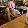 Joshua joined the competition — help win amazing prizes! toddler, child, corn_on_the_cob, high_chair, restaurant, wooden_furniture, smiling, happy, baby_bottle, table, indoor, food, eating, person, face, clothing, hand, seat, light, background