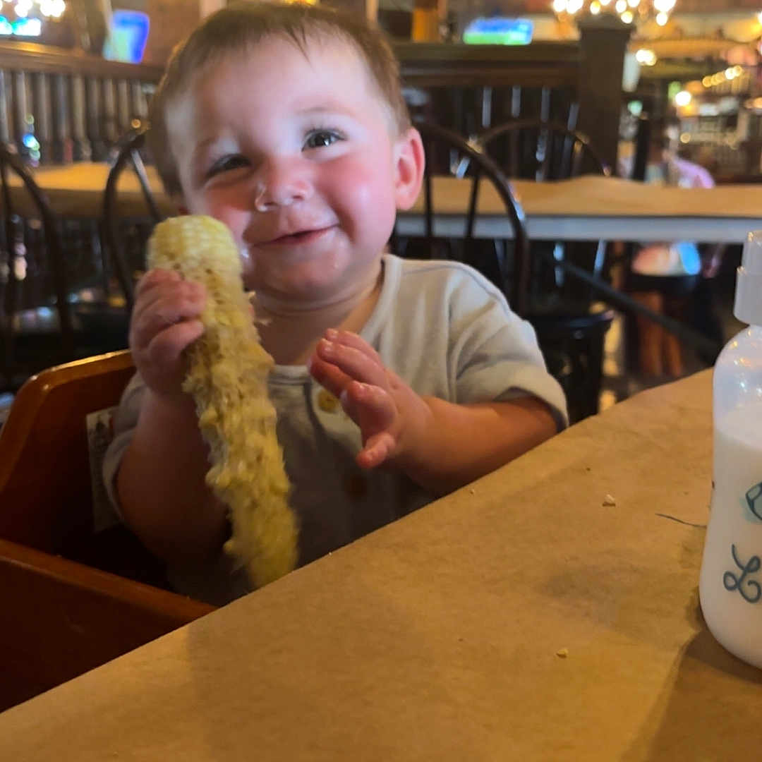 Joshua joined the competition — help win amazing prizes! toddler, child, corn_on_the_cob, high_chair, restaurant, wooden_furniture, smiling, happy, baby_bottle, table, indoor, food, eating, person, face, clothing, hand, seat, light, background