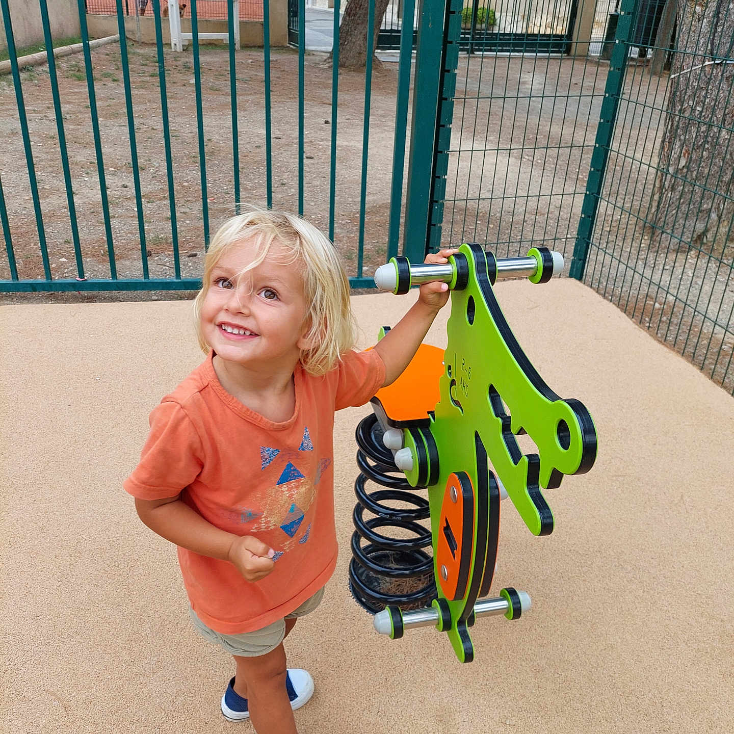 Alina a rejoint le concours — aidez-le/la à gagner de superbes lots ! blond_hair, child, daylight, fence, happy, leisure, orange_shirt, outdoor, park, person, play, playground, recreation, sand, shorts, smiling, sneakers, spring_rider, toddler, tree