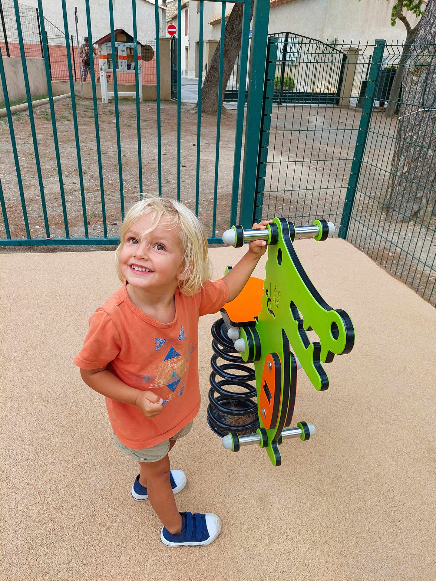 Alina a rejoint le concours — aidez-le/la à gagner de superbes lots ! child, toddler, smiling, playground, spring_rider, outdoor, sand, fence, tree, shorts, sneakers, orange_shirt, blond_hair, happy, play, person, daylight, recreation, leisure, park