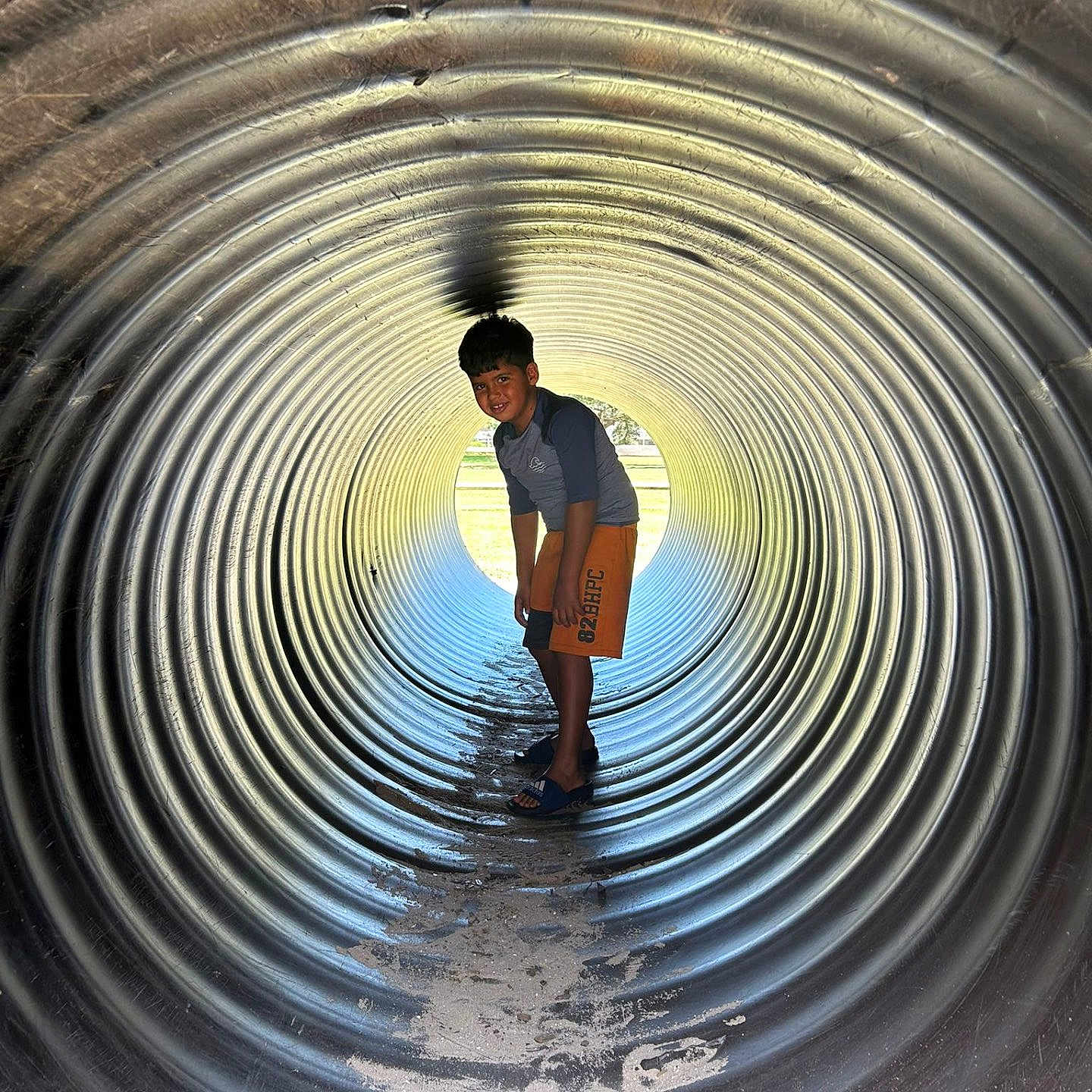 Christian is registered to the contest to win money with this photo: adventure, boy, casual, child, curious, cylindrical, daylight, fun, light, metal, outdoor, playground, sand, shadow, shorts, smiling, standing, texture, tunnel, young