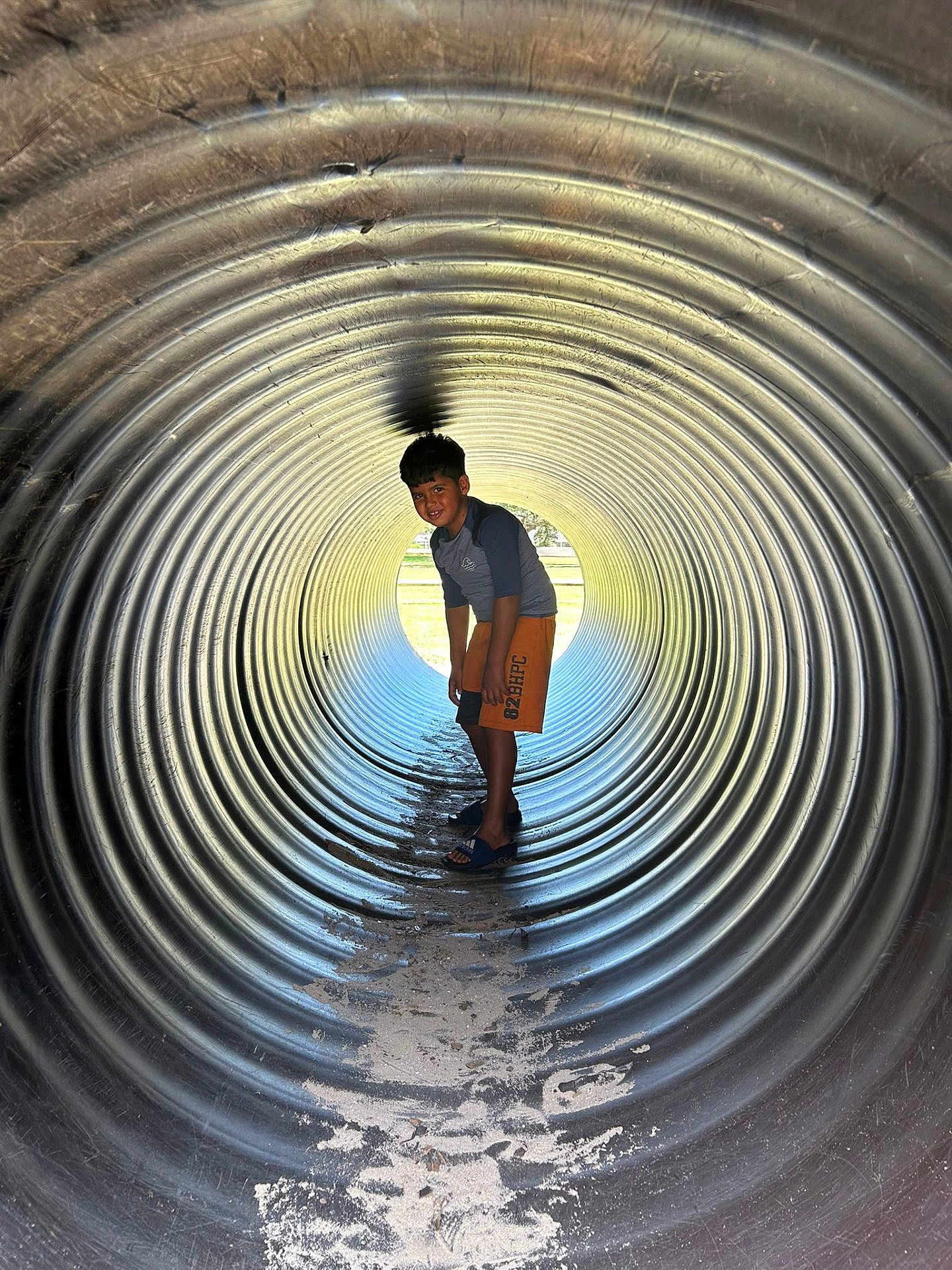 Christian is registered to the contest to win money with this photo: child, boy, tunnel, metal, outdoor, playground, adventure, light, shadow, curious, smiling, shorts, sand, texture, cylindrical, young, standing, casual, fun, daylight