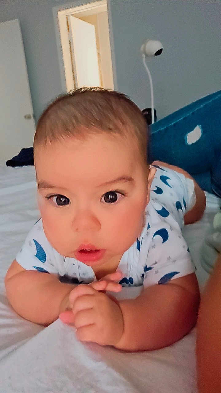 Enzo joined the competition — help win amazing prizes! baby, infant, child, bed, white_bedspread, blue_couch, moon_pattern, curious, wide_eyes, indoor, room, door, propped_up, hands, face, expression, soft_lighting, young_child, casual, home