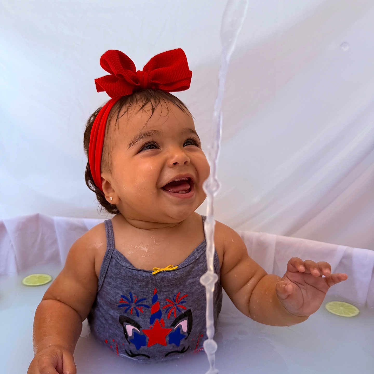Antonella is registered to the contest to win money with this photo: baby, child, red_bow, water, milky_bath, smiling, happy, sleeveless_shirt, face, hand, sitting, lime_slice, water_stream, cute, portrait, infant, headband, skin, playful, indoors