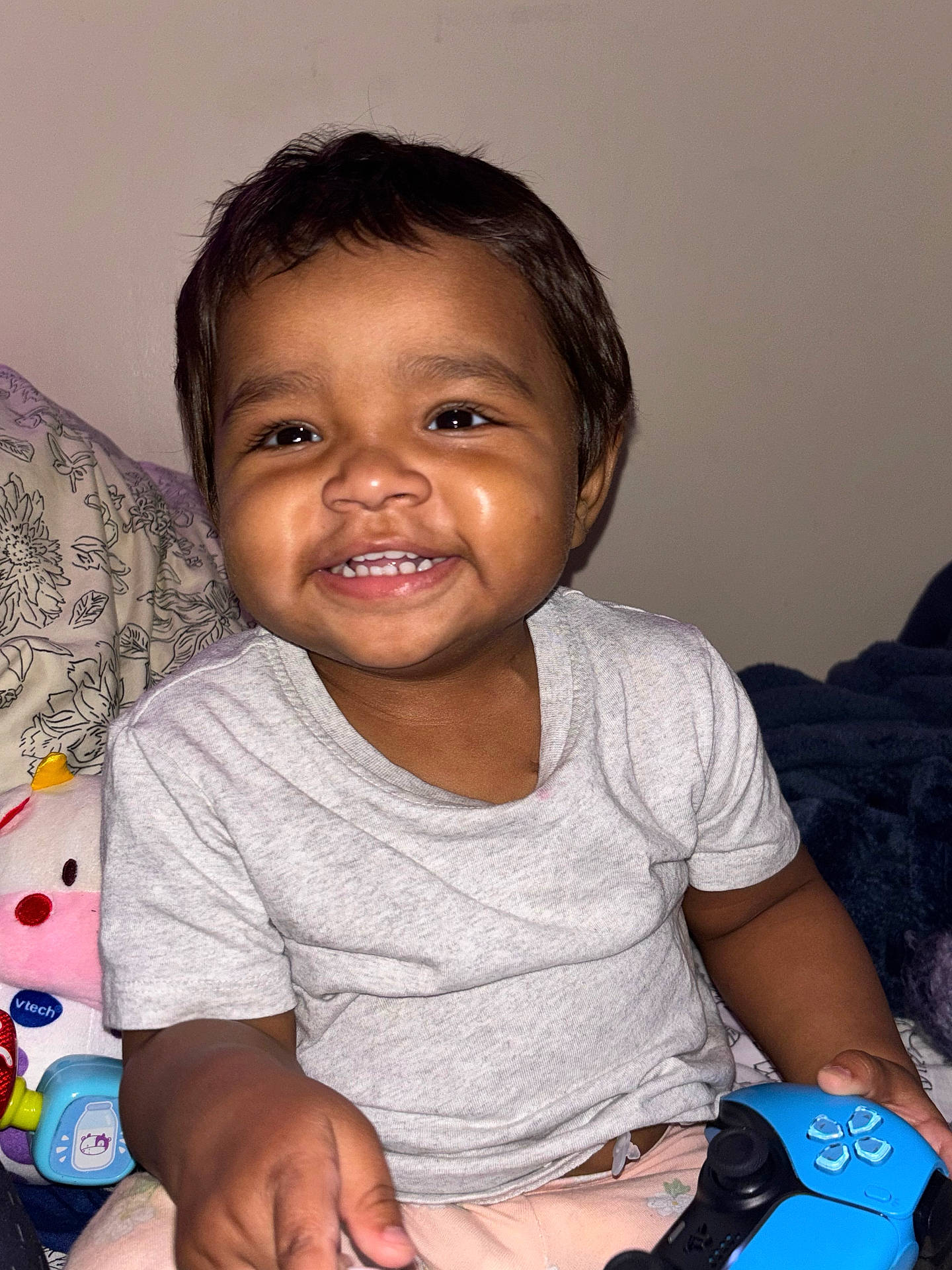 Jamilee joined the competition — help win amazing prizes! bed, blanket, casual_clothing, child, controller, cute, dark_hair, face, hand, happy, indoor, person, playful, plush_toy, shirt, smiling, toddler, toy, video_game, young_child