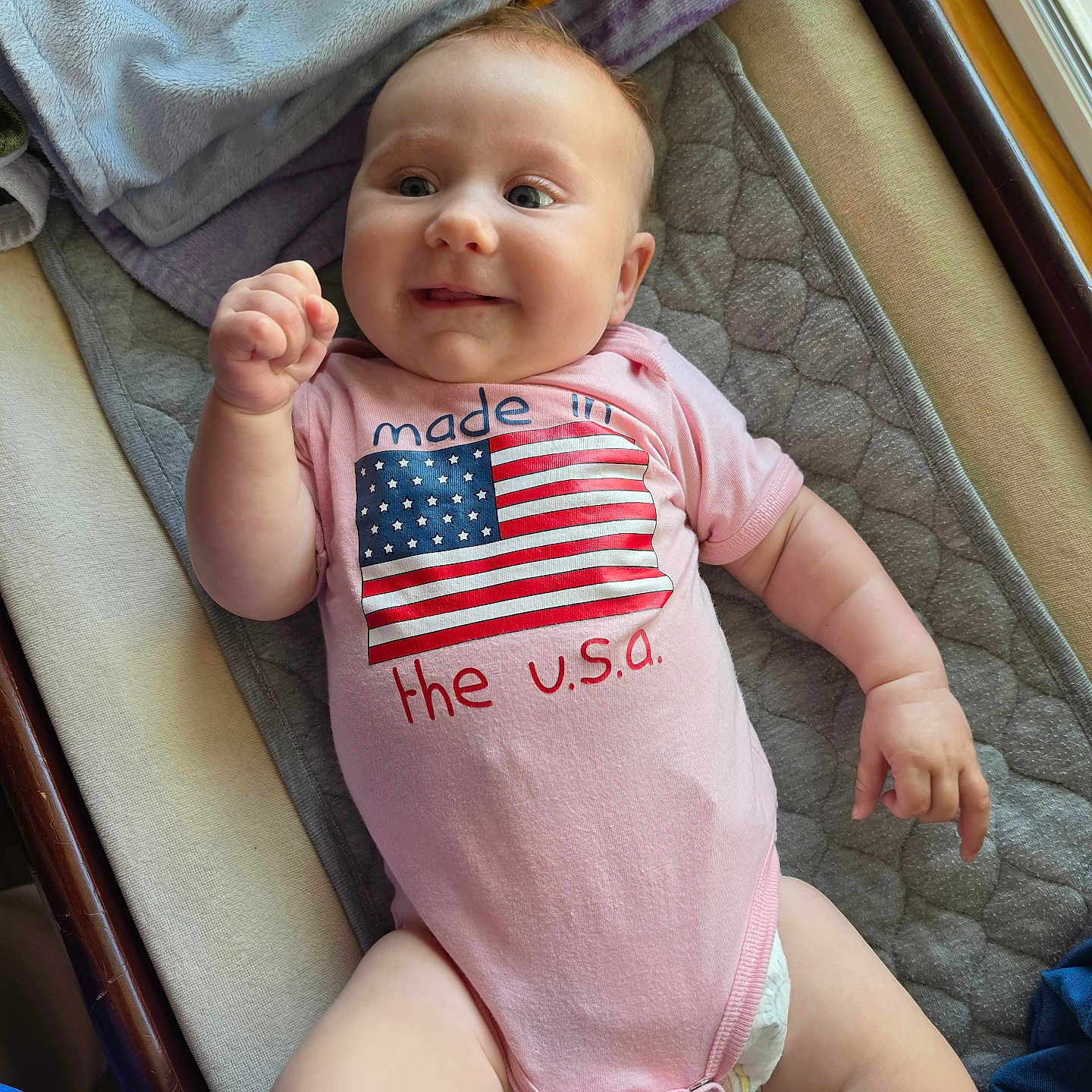 Charli joined the competition — help win amazing prizes! baby, infant, smiling, onesie, american_flag, pink_clothing, lying_down, blanket, diaper, indoors, window, soft_fabric, cute, happy, child, person, skin, face, chubby_arms, natural_light