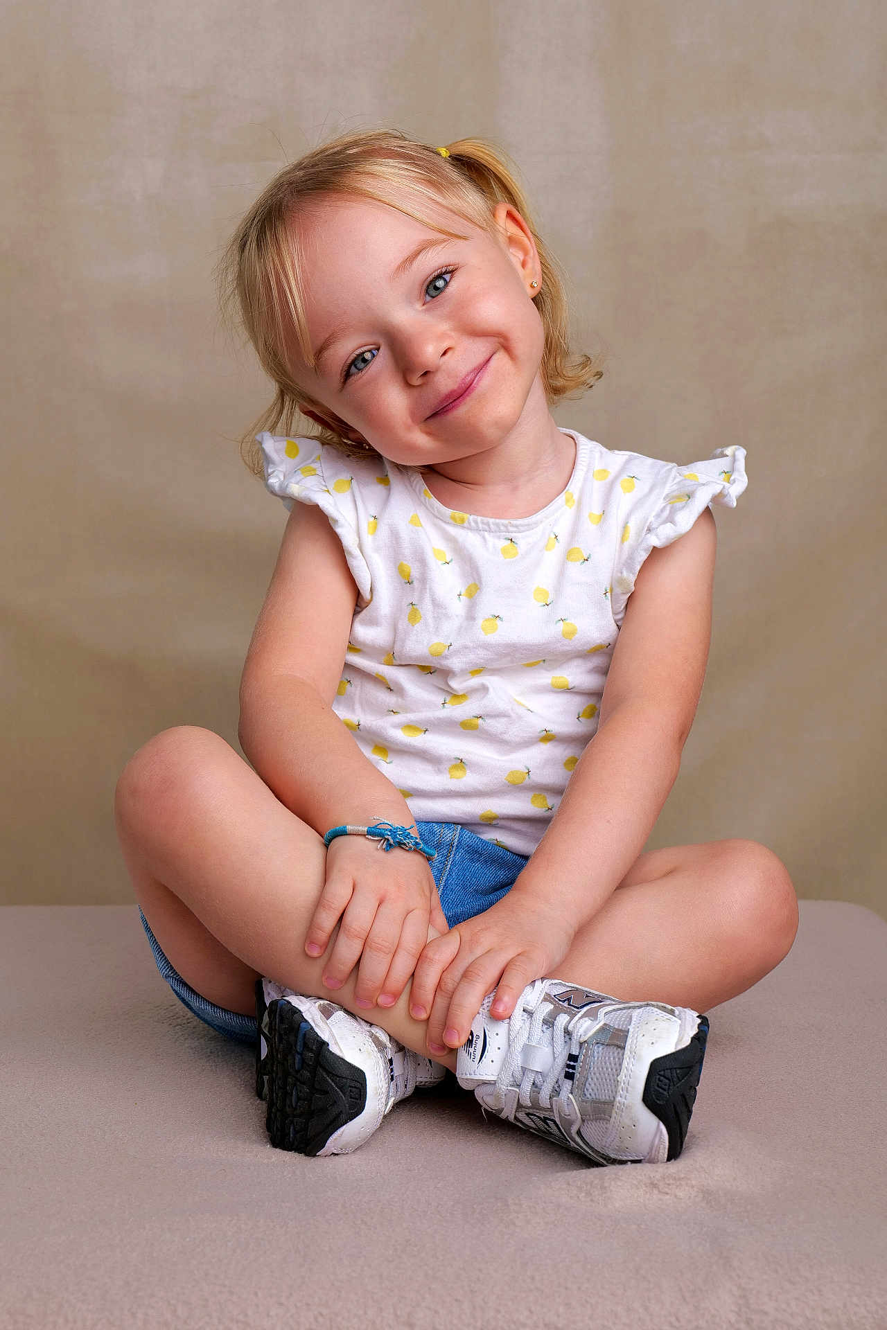 Mencya participe au concours pour gagner de l'argent avec cette photo : background, blonde_hair, bracelet, casual_clothing, child, cross_legged, cute, denim_shorts, girl, happy, innocent, lemon_print, pigtails, portrait, seated, smiling, studio, white_shirt, white_sneakers, young