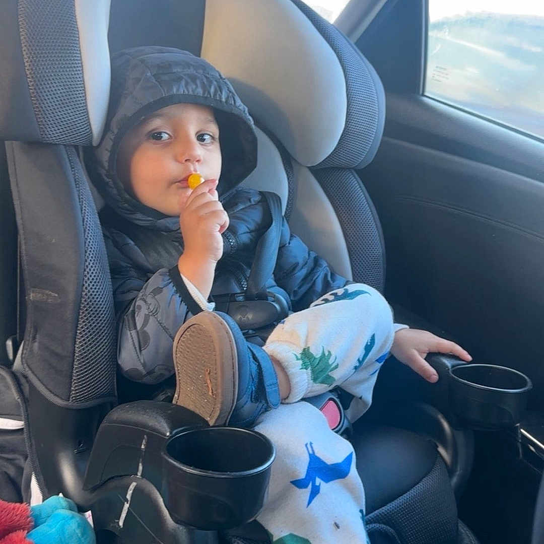 Hamed joined the competition — help win amazing prizes! black_jacket, car_backseat, car_door, car_interior, car_seat, casual_pose, child, child_face, child_in_car, child_relaxing, child_wearing_shoes, cup_holder, dinosaur_pants, hooded_jacket, lollipop, seat_cushion, seatbelt, shoe, toy, window