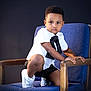 child, boy, chair, blue_cushion, wooden_furniture, white_shirt, striped_shorts, tie, sneakers, portrait, studio_lighting, curly_hair, indoor, fashion, casual, smiling, person, face, looking_at_camera, posing