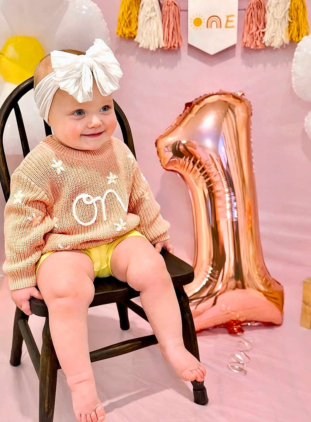 Charleigh is registered to the contest to win money with this photo: baby, toddler, child, bow_headband, white_bow, knitted_sweater, flower_detail, wooden_chair, rose_gold_balloon, number_one_balloon, pink_backdrop, birthday_party, celebration, smiling, sitting, legs, bare_feet, portrait, cute, party_decor