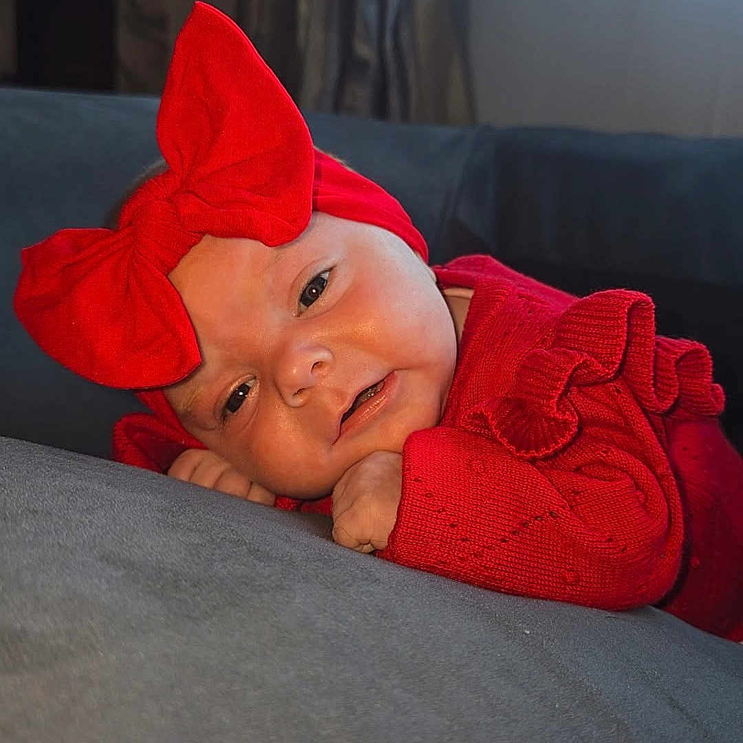 Kyra-Mae is registered to the contest to win money with this photo: baby, child, closeup, cozy, curtains, cute, gray_couch, headwear, home, indoors, infant, natural_light, person, portrait, red_bow, red_clothing, resting, smiling, soft_surface, window