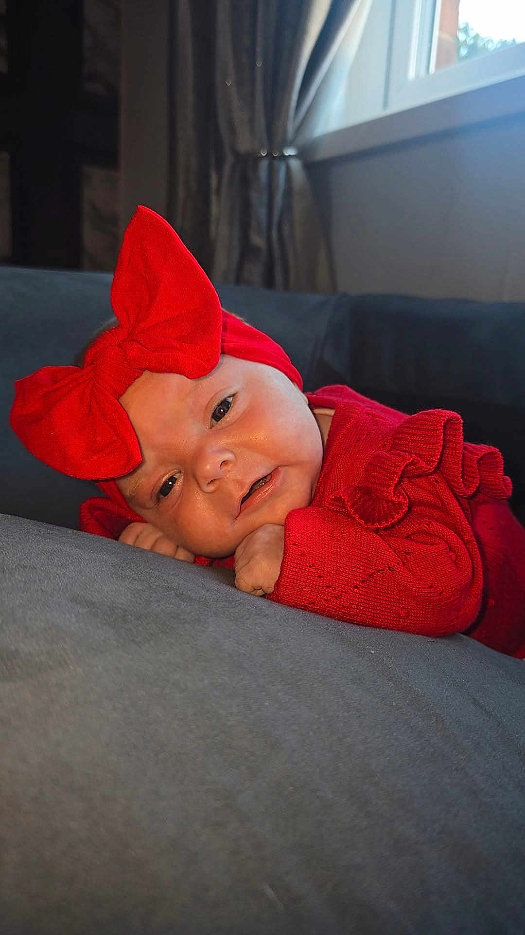 Kyra-Mae is registered to the contest to win money with this photo: baby, infant, red_bow, red_clothing, soft_surface, gray_couch, window, curtains, indoors, portrait, cute, cozy, headwear, closeup, child, smiling, resting, natural_light, home, person