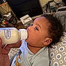 Caspian is registered to the contest to win money with this photo: baby, bottle, milk, curly_hair, infant, child, feeding, pillow, pattern, indoor, person, cute, clothing, blue_shirt, face, hand, lying_down, room, furniture, background