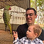 child, man, parrot, bird, cage, wire_mesh, outdoor, trees, nature, glasses, plaid_shirt, curious, smile, holding, adult, baby, wildlife, daylight, interaction, family