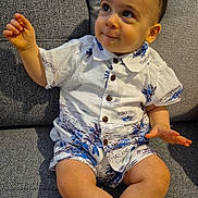 Amir a rejoint le concours — aidez-le/la à gagner de superbes lots ! toddler, child, couch, clothing, shirt, shorts, face, legs, feet, sitting, indoor, looking_up, curious, smile, buttoned_shirt, patterned_clothing, skin, hair, hand, furniture