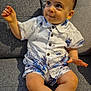toddler, child, couch, clothing, shirt, shorts, face, legs, feet, sitting, indoor, looking_up, curious, smile, buttoned_shirt, patterned_clothing, skin, hair, hand, furniture