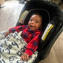 baby, bag, black_car_seat, blanket, car_seat, child, cushion, face, floor, footwear, hand, head, home, indoor, infant, person, plaid, red_clothing, smiling, wooden_floor