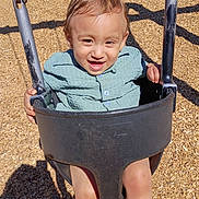 Rene Bolcao is registered to the contest to win money with this photo: toddler, child, playground, swing, smiling, happy, outdoor, portrait, playground_equipment, wood_chips, sunlight, shadow, chains, seated, blue_shirt, brown_hair, face, hands, legs, joy