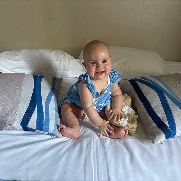 Leïa participe au concours pour gagner de l'argent avec cette photo : baby, barefoot, bed, blue_outfit, child, cute, furniture, happy, hat, headboard, indoor, infant, person, pillow, plush, portrait, sitting, smiling, striped_pillow, white_bed_sheet