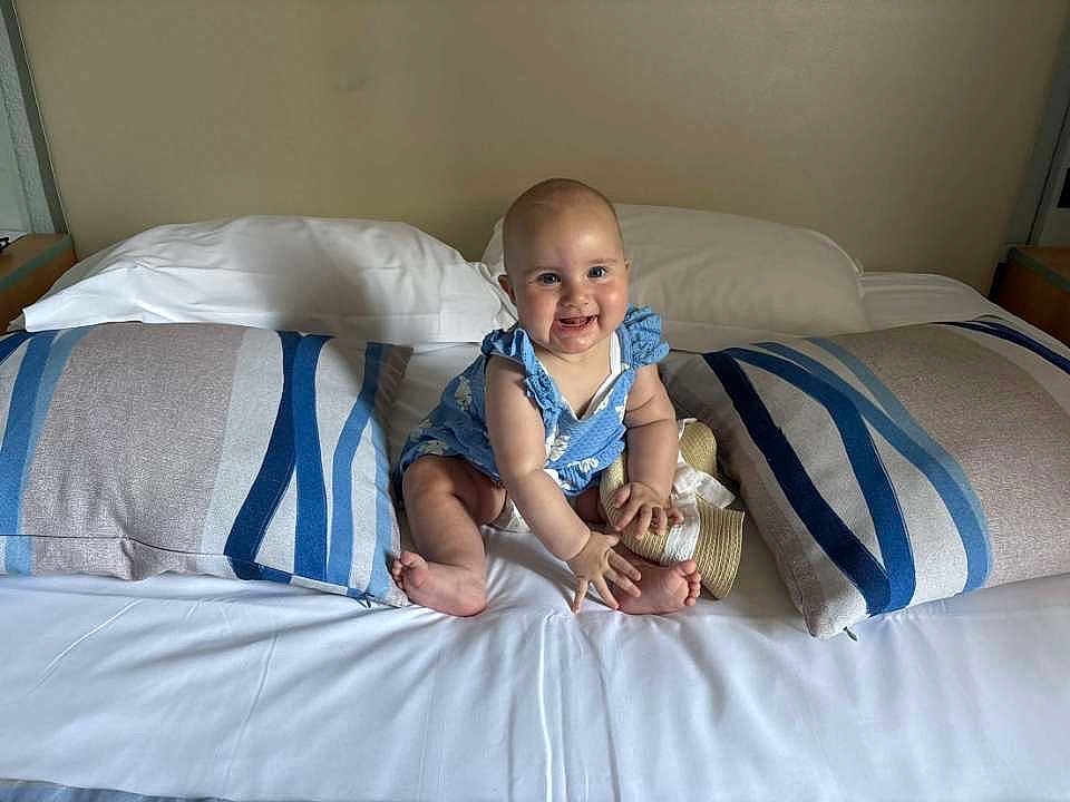 Leïa participe au concours pour gagner de l'argent avec cette photo : baby, child, bed, pillow, striped_pillow, blue_outfit, hat, smiling, indoor, headboard, white_bed_sheet, barefoot, infant, sitting, cute, happy, person, portrait, plush, furniture