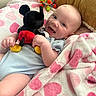 baby, smiling, plush_toy, mickey_mouse, blanket, heart_pattern, couch, toy, infant, happy, child, cute, soft_fabric, indoor, cozy, person, lying_down, colorful, playful, comfort