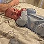 baby, infant, bed, bedding, pacifier, foot, adult, onesie, smile, happy, lying_down, indoor, blanket, remote_control, fabric, person, skin, hand, face, room