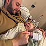 baby, beard, bed, blanket, casual_clothing, comfort, couch, face, family, hand, holding_hands, hoodie, indoor, love, man, person, pillow, rest, skin, sleepy
