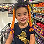 child, smile, shopping_cart, grocery_store, hat, witch_hat, braids, orange_hair, purple_ribbons, unicorn_shirt, necklace, snacks, candy, aisle, happy, portrait, retail, store_shelf, cute, fashion