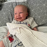 Tadhg is registered to the contest to win money with this photo: baby, smiling, bed, pillow, blanket, grey, geometric_pattern, clothing, red_crab, infant, cute, happy, face, head, resting, soft_texture, comfort, indoors, person, portrait