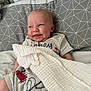 baby, smiling, bed, pillow, blanket, grey, geometric_pattern, clothing, red_crab, infant, cute, happy, face, head, resting, soft_texture, comfort, indoors, person, portrait