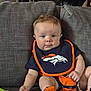 baby, infant, child, couch, bib, orange, blue, clothing, footwear, socks, person, face, eyes, hair, hand, seated, indoor, furniture, sports_logo, expression