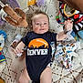 baby, infant, onesie, denver, football, plush_toy, blanket, patterned_blanket, colorful, smiling, reaching, lying_down, baby_accessories, cute, child, indoors, soft_toys, blue_clothing, orange_logo, happy