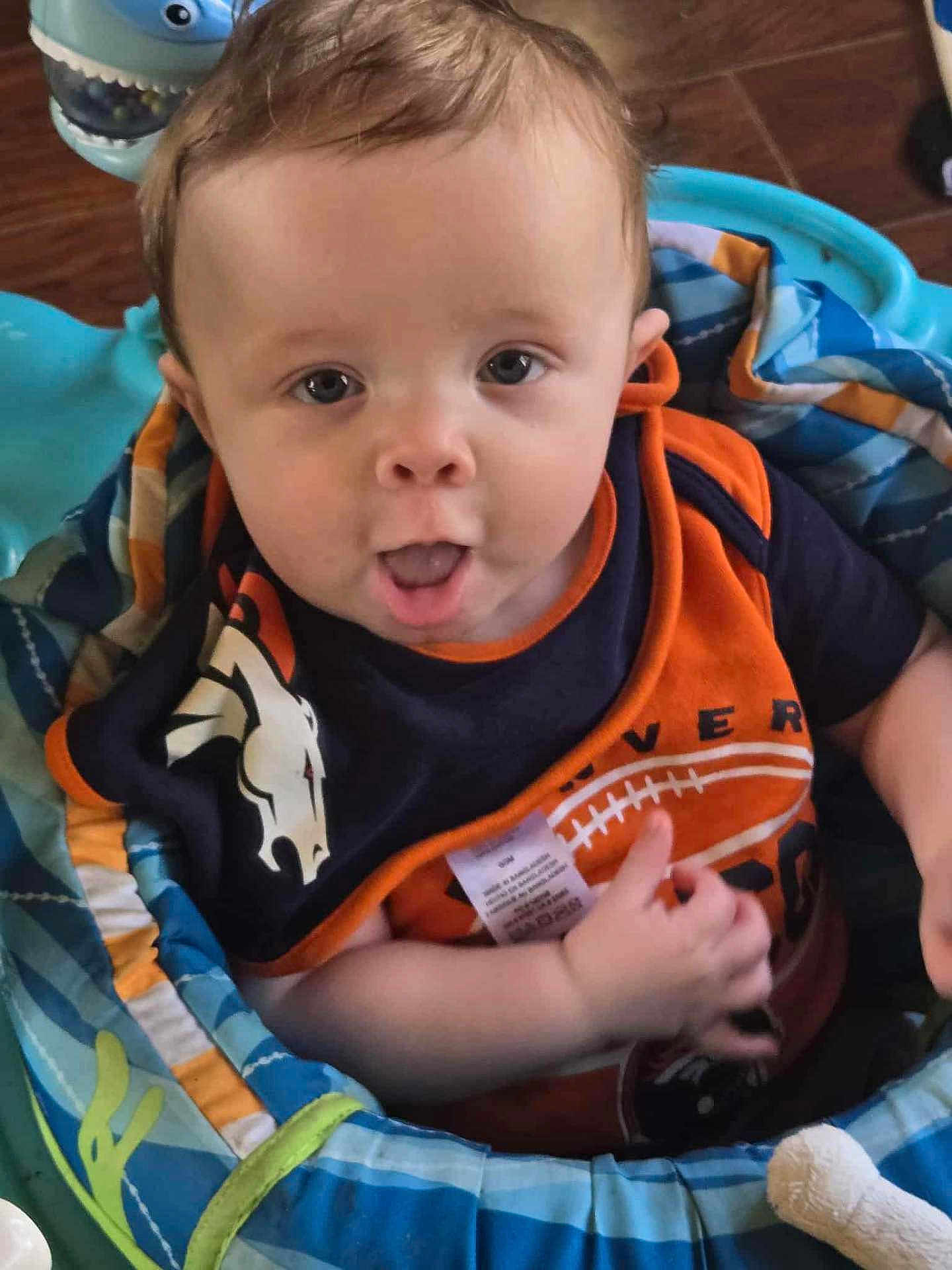 Isiah joined the competition — help win amazing prizes! baby, bib, blue, child, clothing, curious, eyes, face, floor, indoor, infant, mouth, navy, orange, padded_seat, person, playful, seat, toy, wooden_floor