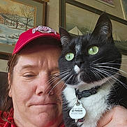 Diamond Marie joined the competition — help win amazing prizes! person, cat, indoor, selfie, red_hat, black_and_white_cat, collar, pet_tag, green_eyes, hand, painting, wall_art, framed_picture, casual_clothing, close_up, facial_expression, whiskers, home_interior, portrait, furniture