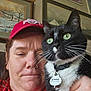 person, cat, indoor, selfie, red_hat, black_and_white_cat, collar, pet_tag, green_eyes, hand, painting, wall_art, framed_picture, casual_clothing, close_up, facial_expression, whiskers, home_interior, portrait, furniture