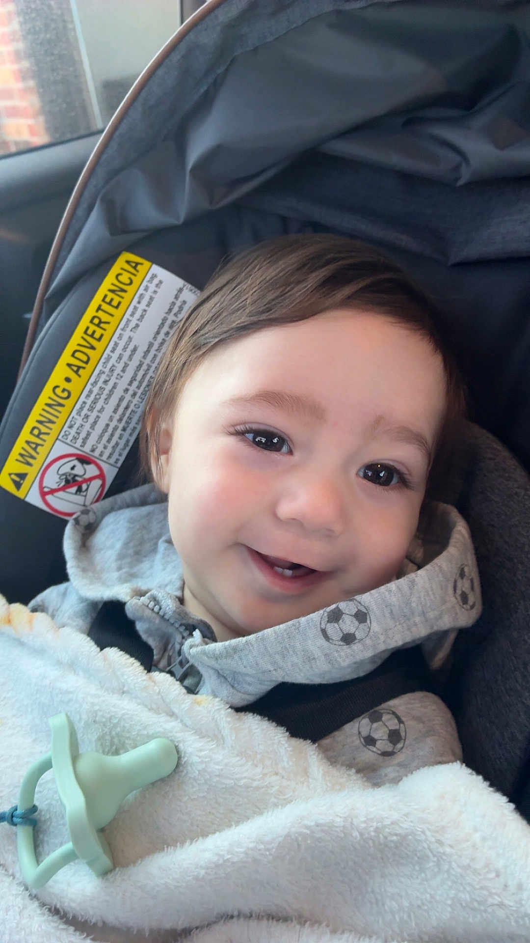 Oliver joined the competition — help win amazing prizes! baby, child, infant, smile, teeth, car_seat, pacifier, blanket, hoodie, soccer_pattern, safety_warning, strap, close_up, portrait, eyes, hair, window, cozy, happy, seat