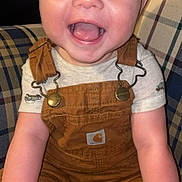 Levi is registered to the contest to win money with this photo: baby, infant, smiling, happy, child, overalls, brown_clothing, plaid_chair, patterned_fabric, sitting, cute, portrait, indoors, person, young_child, face, cheerful, skin, clothing, hands