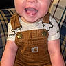 baby, infant, smiling, happy, child, overalls, brown_clothing, plaid_chair, patterned_fabric, sitting, cute, portrait, indoors, person, young_child, face, cheerful, skin, clothing, hands