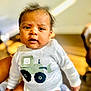 Sri Thanvik is registered to the contest to win money with this photo: baby, infant, child, onesie, clothing, face, eyes, hair, expression, indoors, blurred_background, person, skin, sitting, cute, adorable, young, portrait, smiling, bright