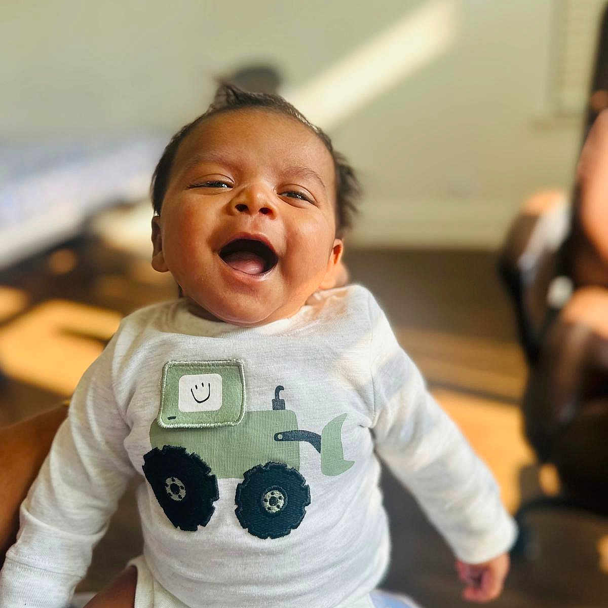 Sri Thanvik joined the competition — help win amazing prizes! baby, blurred_background, cheerful, child, clothing, cute, face, happy, human, indoor, infant, joyful, onesie, person, portrait, sitting, smiling, sunlight, tractor_design, warm_light