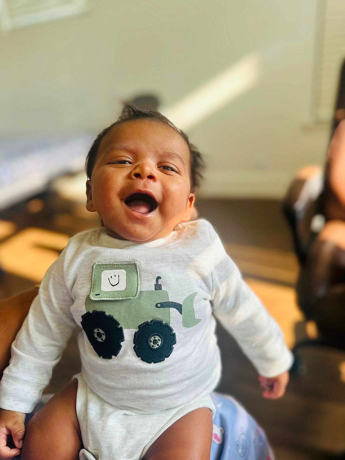 Sri Thanvik joined the competition — help win amazing prizes! baby, child, smiling, happy, onesie, clothing, indoor, sunlight, face, person, infant, cute, joyful, portrait, sitting, human, cheerful, warm_light, blurred_background, tractor_design