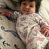 baby, bed, blanket, child, cozy, curly_hair, cute, dinosaur_print, happy, indoors, infant, lying_down, onesie, person, pillow, portrait, sleepwear, smiling, soft_lighting, toddler