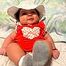 baby, blanket, child, chubby_cheeks, cute, floral_pattern, footwear, happy, hat, indoors, infant, lying_down, person, playful, polka_dots, portrait, red_clothing, smiling, socks, white_hat